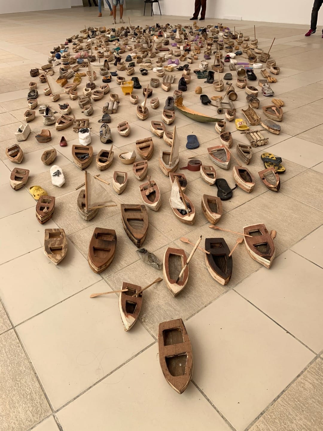 Art installation featuring numerous small wooden boats arranged on a tiled floor in a gallery setting.