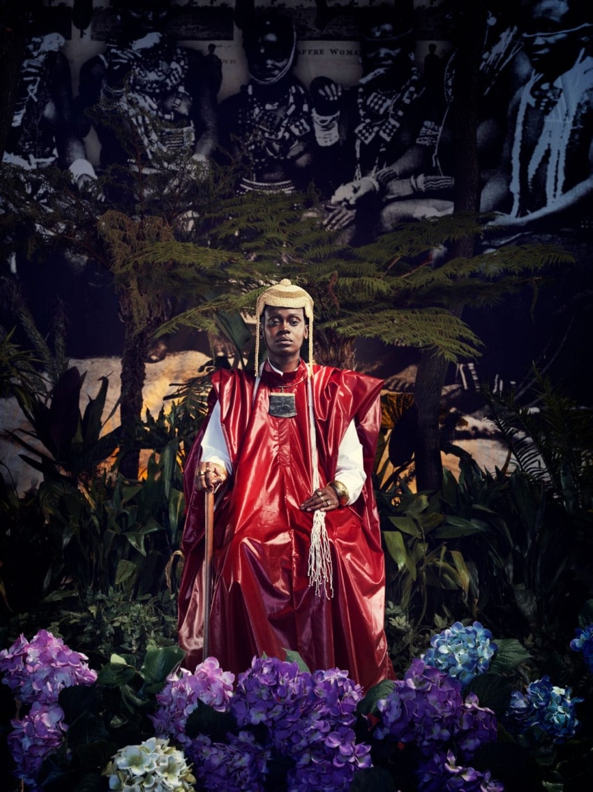Person in red ceremonial attire sits among vibrant flowers and greenery with a dark, patterned background.