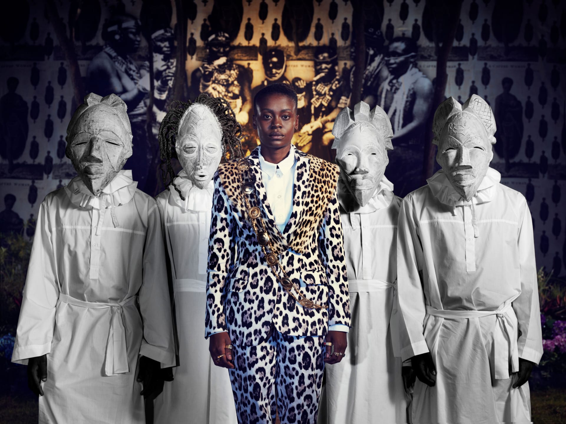 Person in leopard print suit stands with four individuals in white traditional masks and robes.