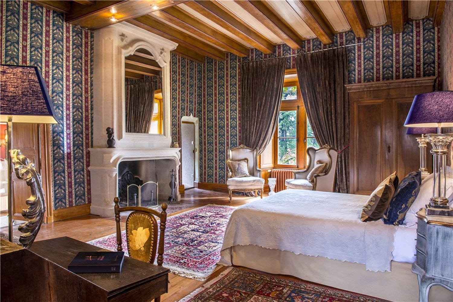 Luxurious bedroom with ornate furniture, vintage wallpaper, a cozy fireplace, and large windows with elegant drapes.