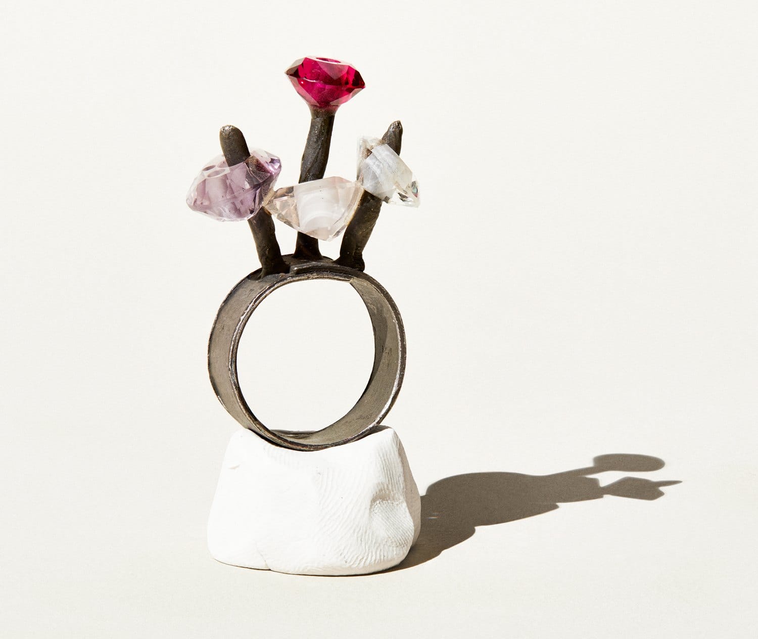 Silver ring with three branches, each holding a differently colored gemstone, displayed on a white geometric stand.