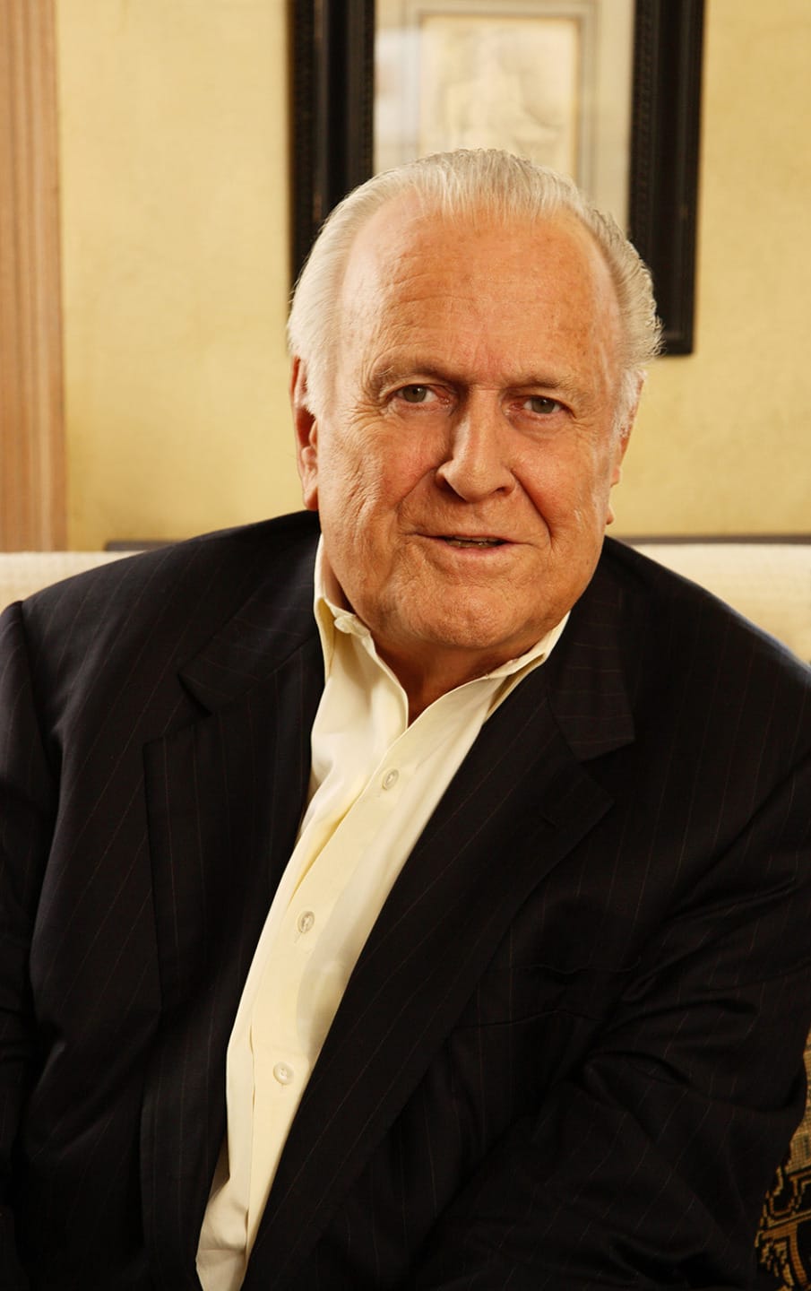 Elderly man in a suit seated on a couch, smiling warmly with a framed picture in the background.