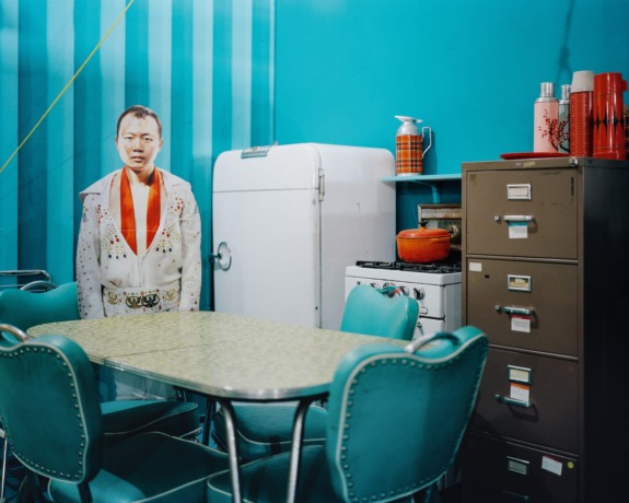 Retro kitchen with teal chairs, vintage refrigerator, filing cabinet, and Elvis Presley cardboard cutout in a blue room.