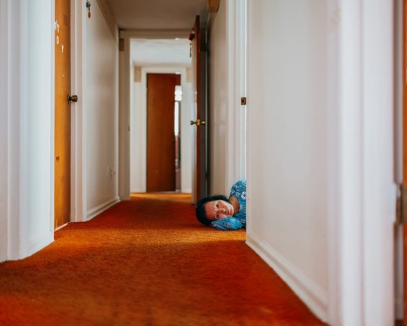 Person lying on orange carpeted hallway floor, peeking around a corner with a neutral expression.