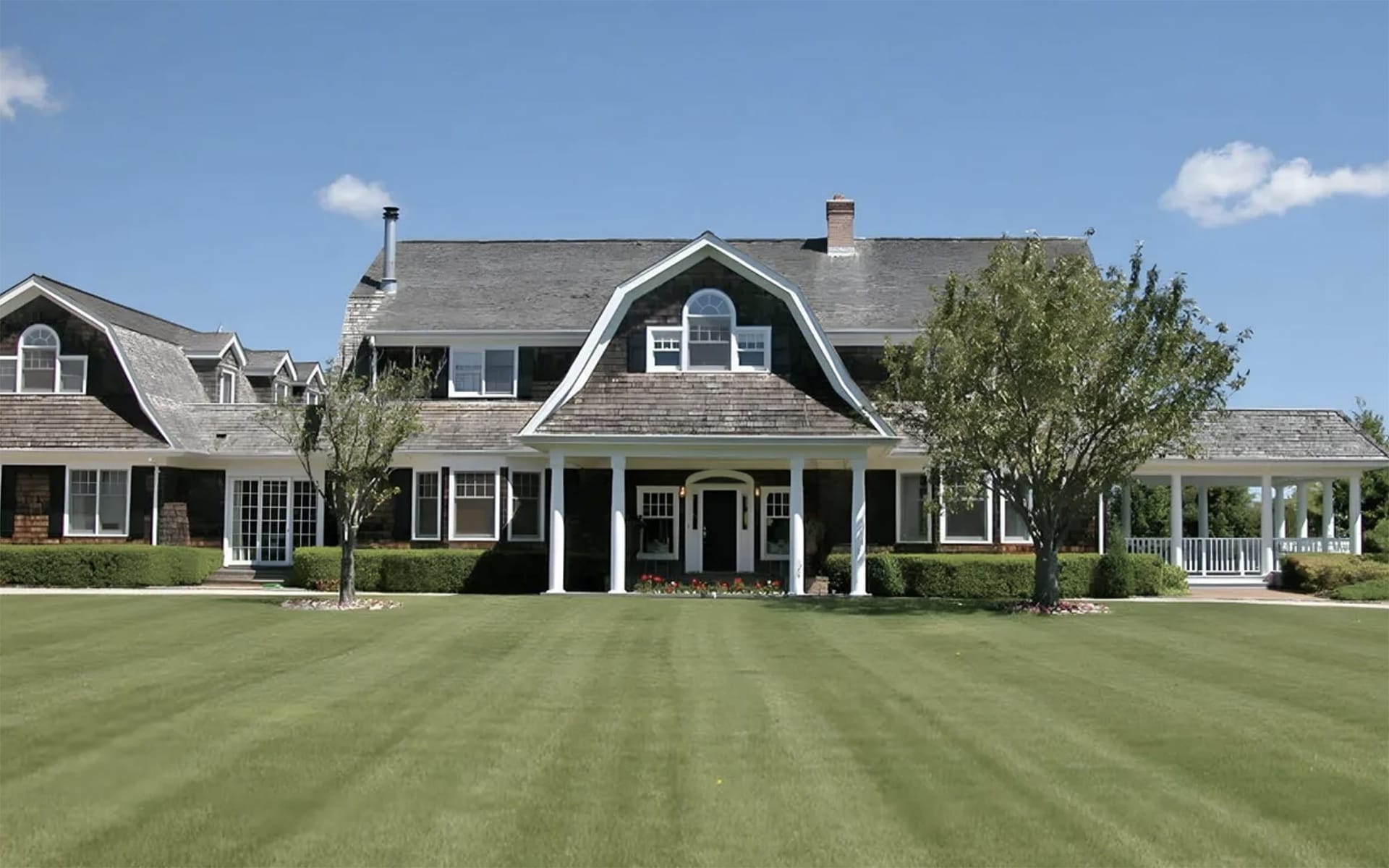 Large two-story house with gray roof, white-trimmed windows, and columns, surrounded by green lawn and trees under a blue sky.