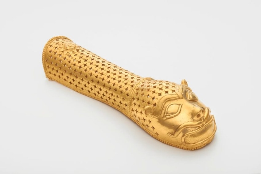 Golden ceremonial object with intricate patterns and a sculpted face on one end, displayed against a plain background.