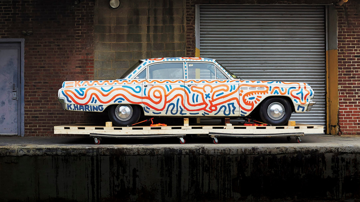 Vintage car with colorful, abstract patterns by artist, displayed on pallets in an industrial setting.
