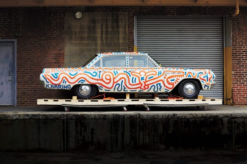 Vintage car painted with colorful abstract patterns parked in front of a brick building with a metal garage door.