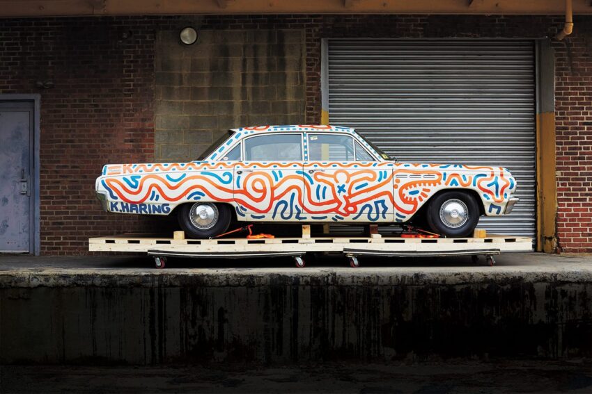Vintage car painted with colorful abstract patterns parked in front of a brick building with a metal garage door.