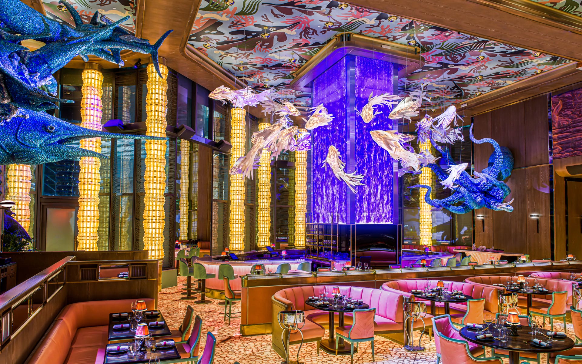 Vibrant restaurant interior with colorful fish sculptures, patterned ceiling, blue lighting, and elegantly set dining tables.