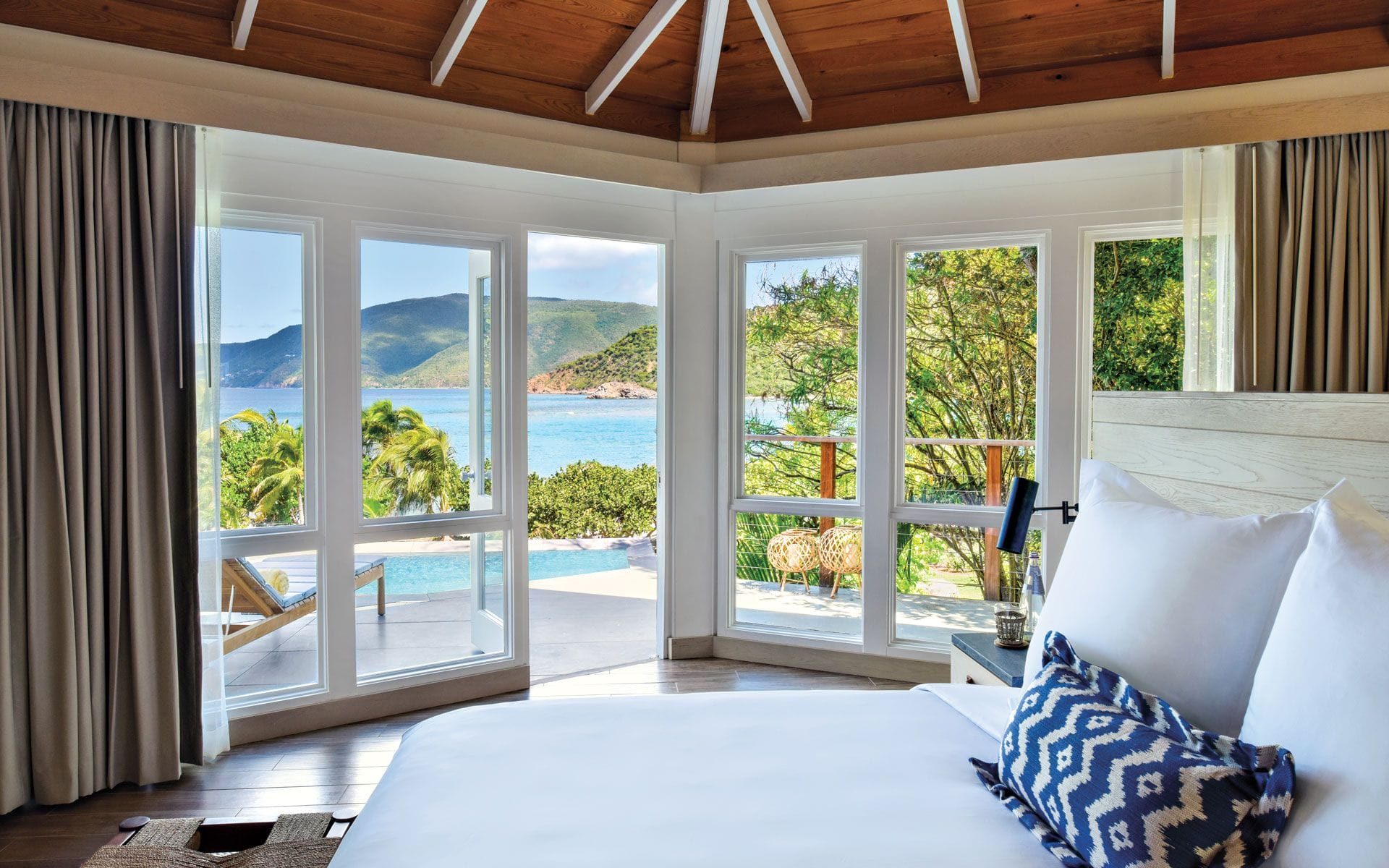 Scenic view from a cozy bedroom featuring large windows overlooking a blue ocean and lush green hills.