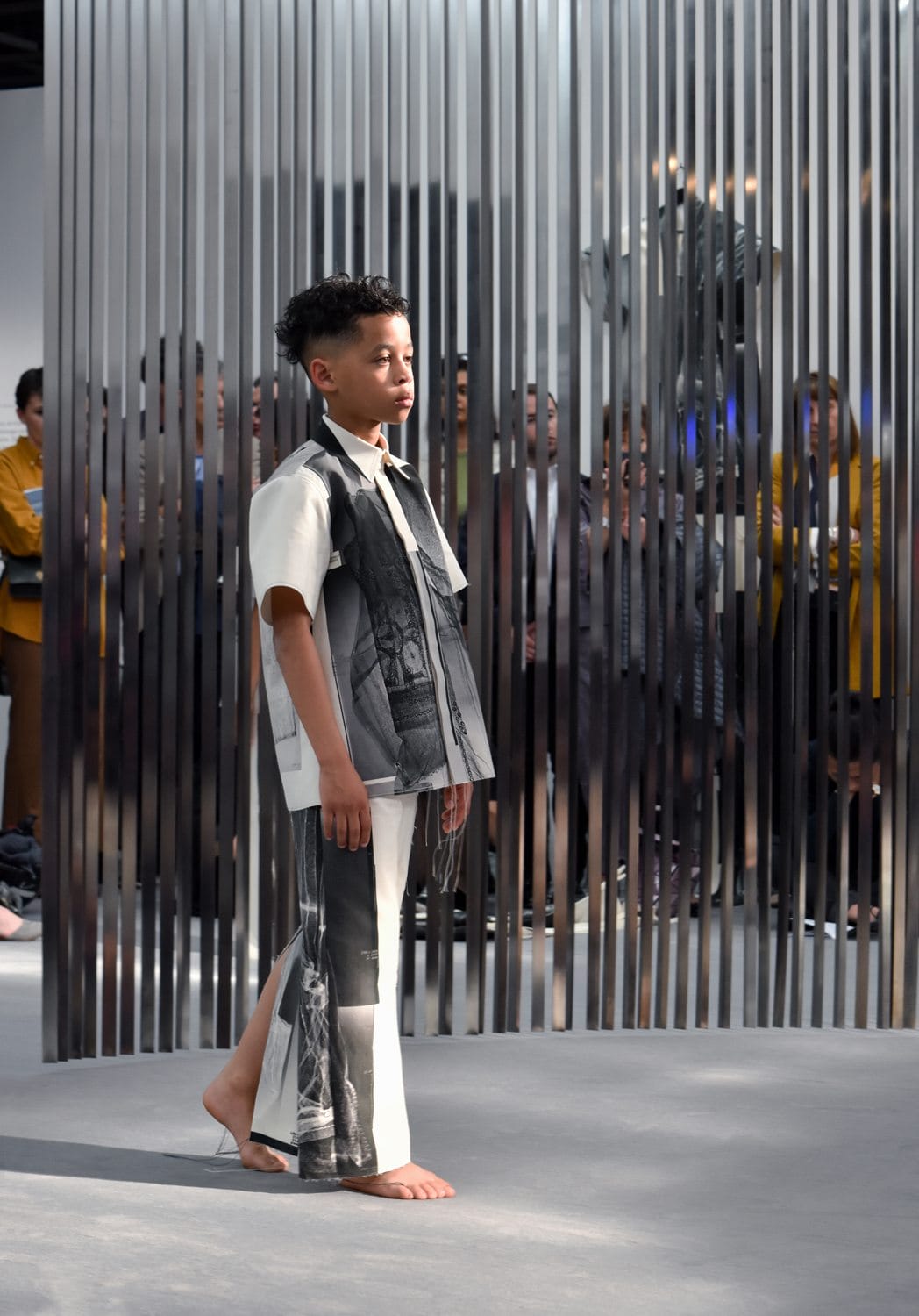 Child model in black and white patterned outfit walks barefoot on runway with metallic backdrop and spectators behind.