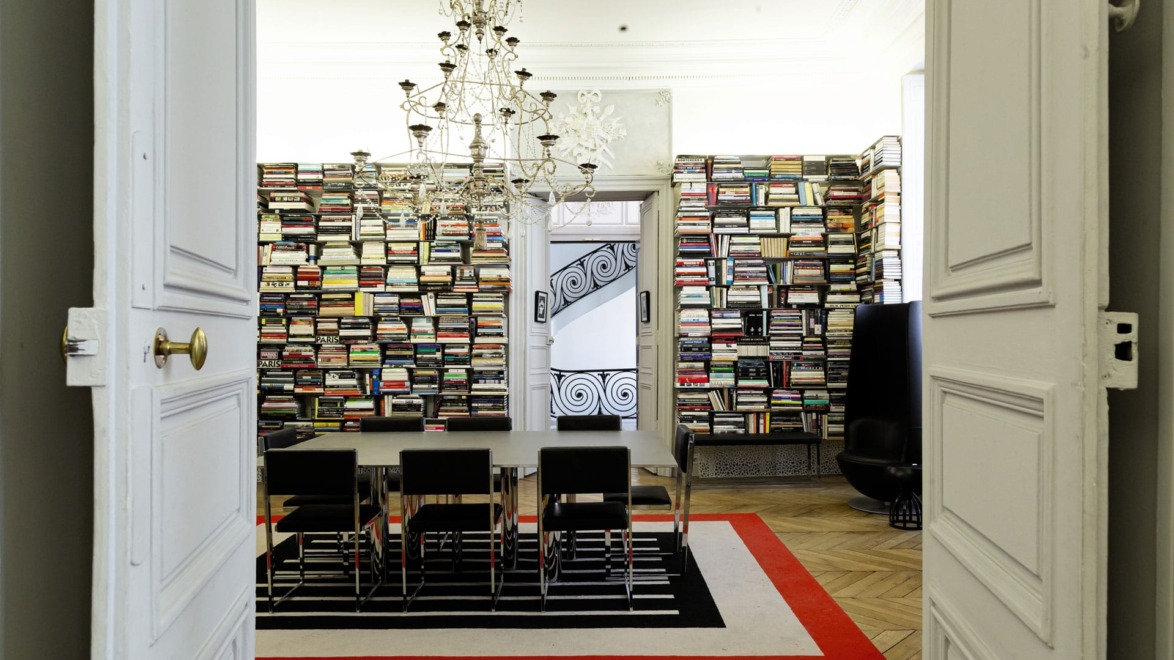Dining room with a chandelier, chairs, and walls filled with books, visible through open double doors.