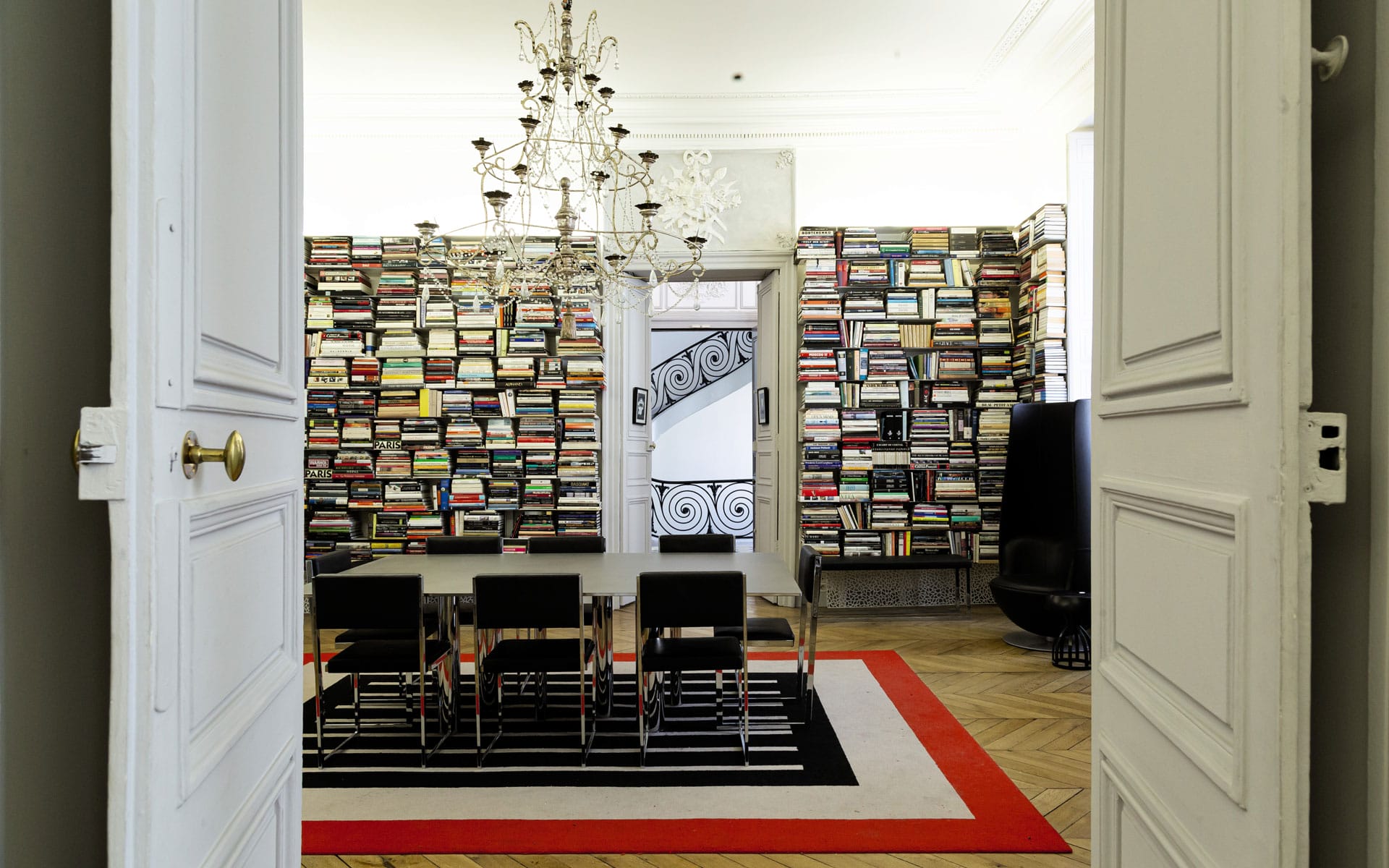 Dining room with a chandelier, chairs, and walls filled with books, visible through open double doors.