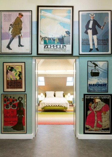 Colorful vintage posters decorate the walls of a room with a view into a modern bedroom featuring a neatly made bed.