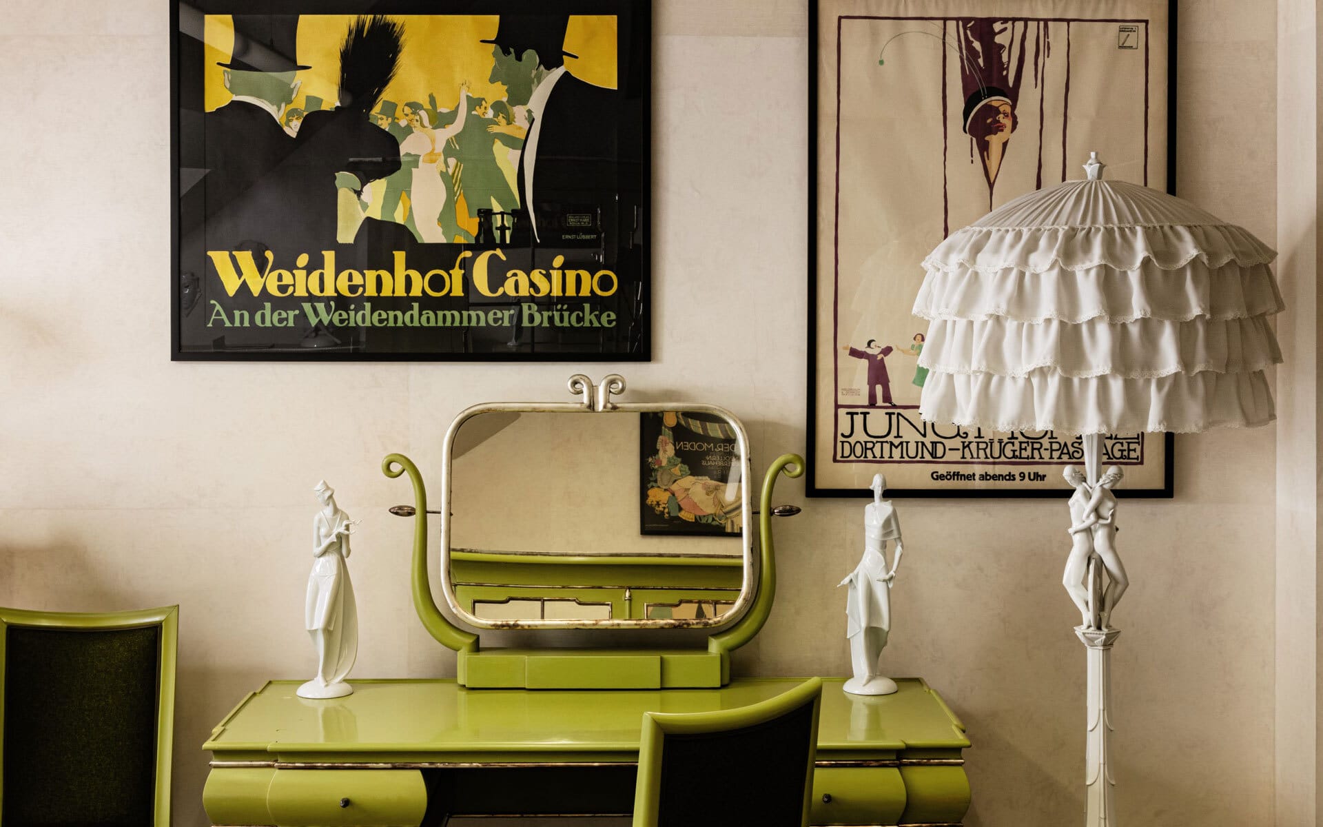 Art deco vanity with green dresser, ornate mirror, period lamps, and vintage posters on wall in a stylish interior setting.