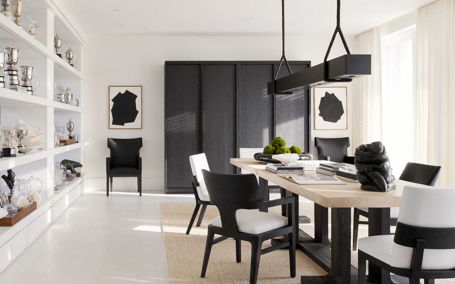 Modern dining room with a wooden table, black and white chairs, large pendant light, and trophy shelves on the left.
