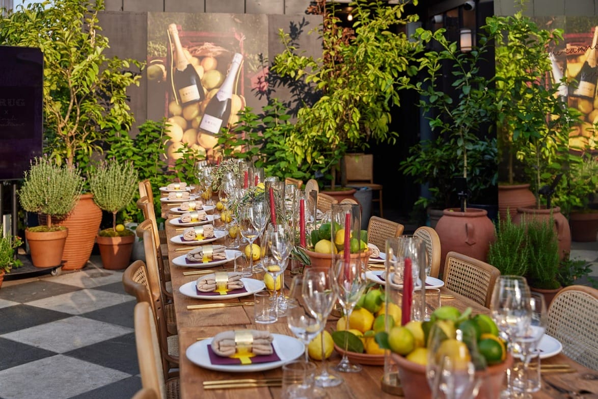 Outdoor dining table with place settings, glasses, fruit bowls, and wine, surrounded by lush green plants and posters.
