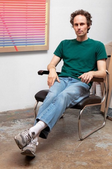 Person sitting on a chair in a casual setting, wearing a green shirt and jeans, with artwork in the background.