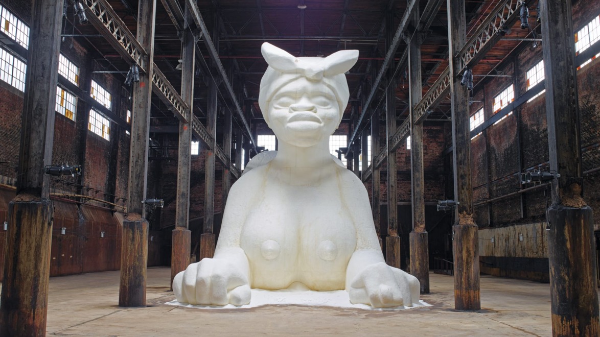 Giant white sculpture of a woman with a headscarf, displayed in an industrial warehouse setting with wooden beams.