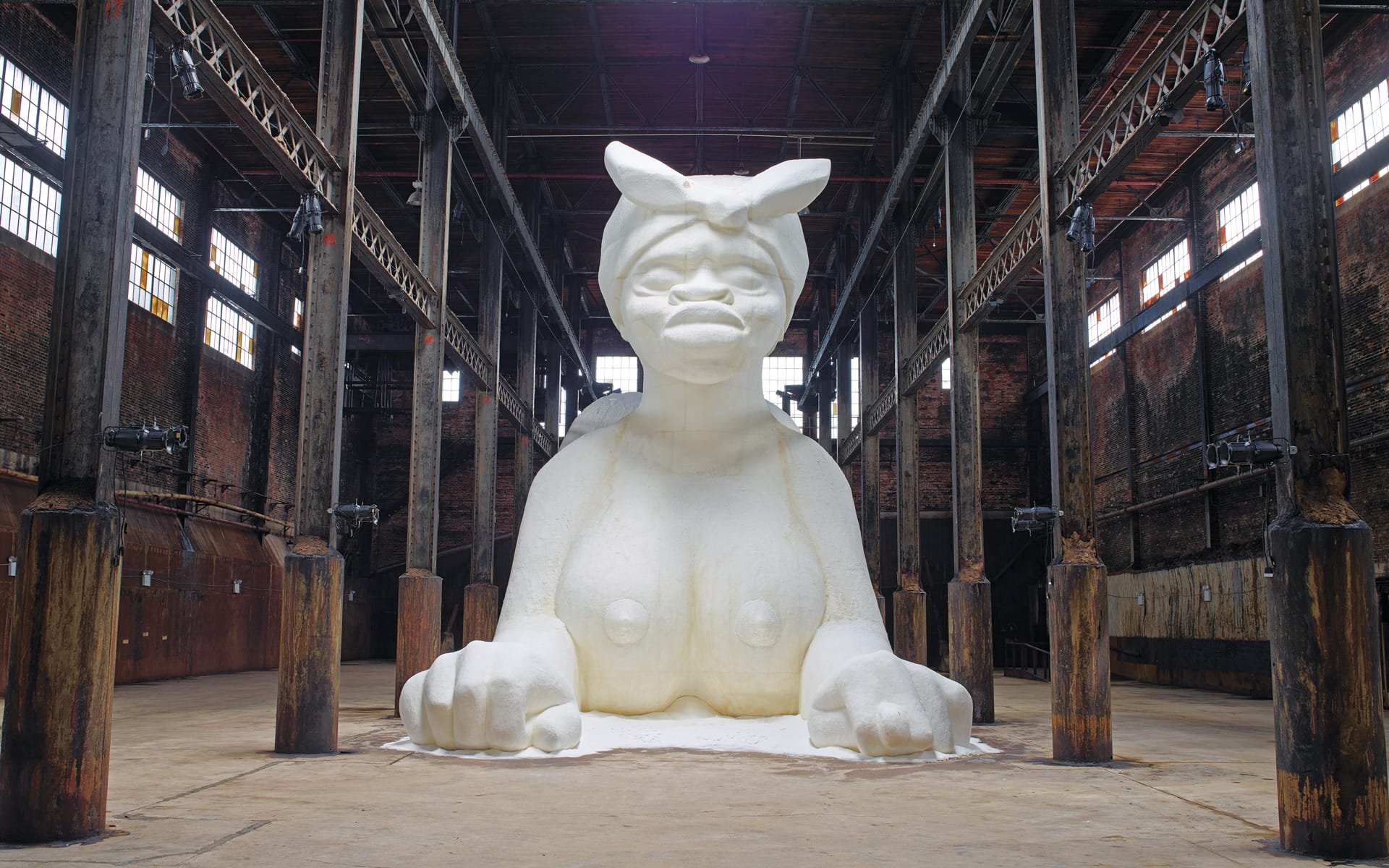 Giant white sculpture of a woman with a headscarf, displayed in an industrial warehouse setting with wooden beams.