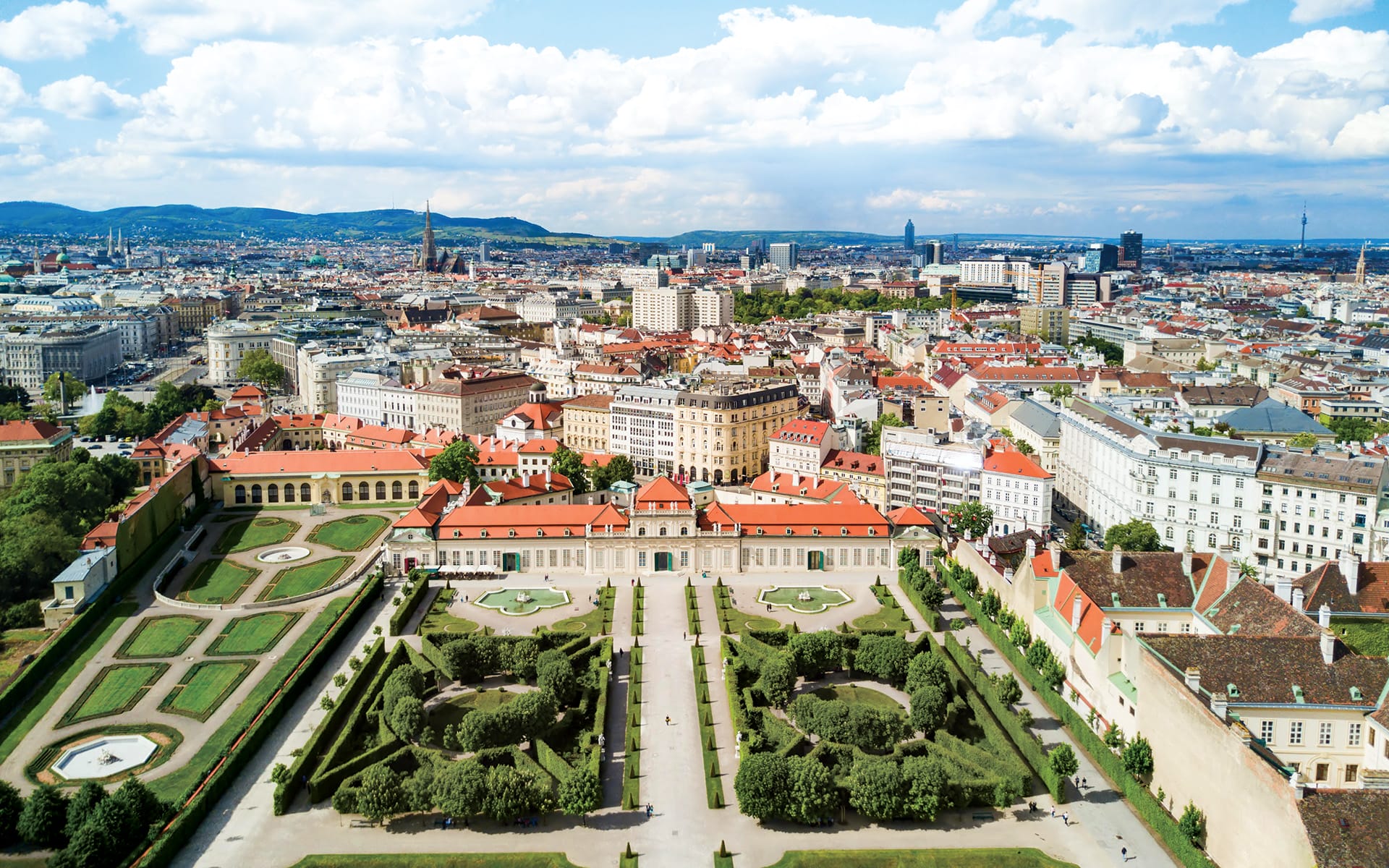 Aerial view of a cityscape with historic and modern buildings, manicured gardens, and a distant mountain backdrop on a clear day.