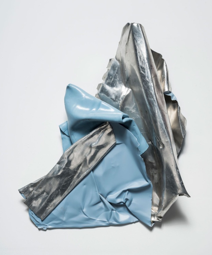 Abstract sculpture with crumpled blue and silver metallic-like sheets on a neutral background.