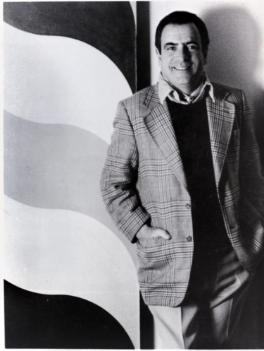 Man in a plaid blazer smiles while standing next to a wavy patterned wall, hands in pockets, black and white photo.