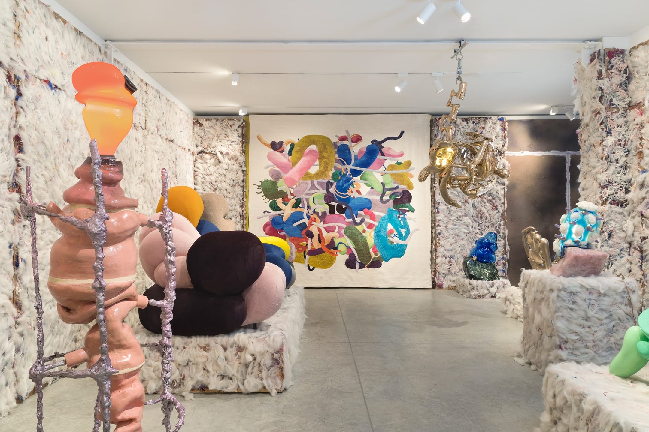 Contemporary art gallery exhibition with colorful abstract sculptures and a vibrant textile piece on the wall.