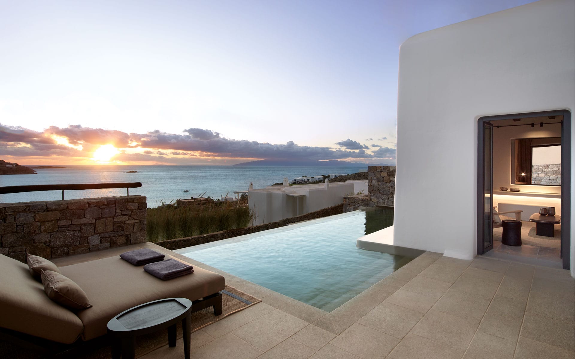 Infinity pool with lounge chair overlooking the ocean at sunset, modern architecture with open room view.