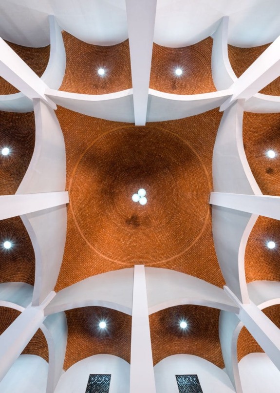 View of a geometric ceiling with white beams and a central brick dome, illuminated by circular light fixtures.