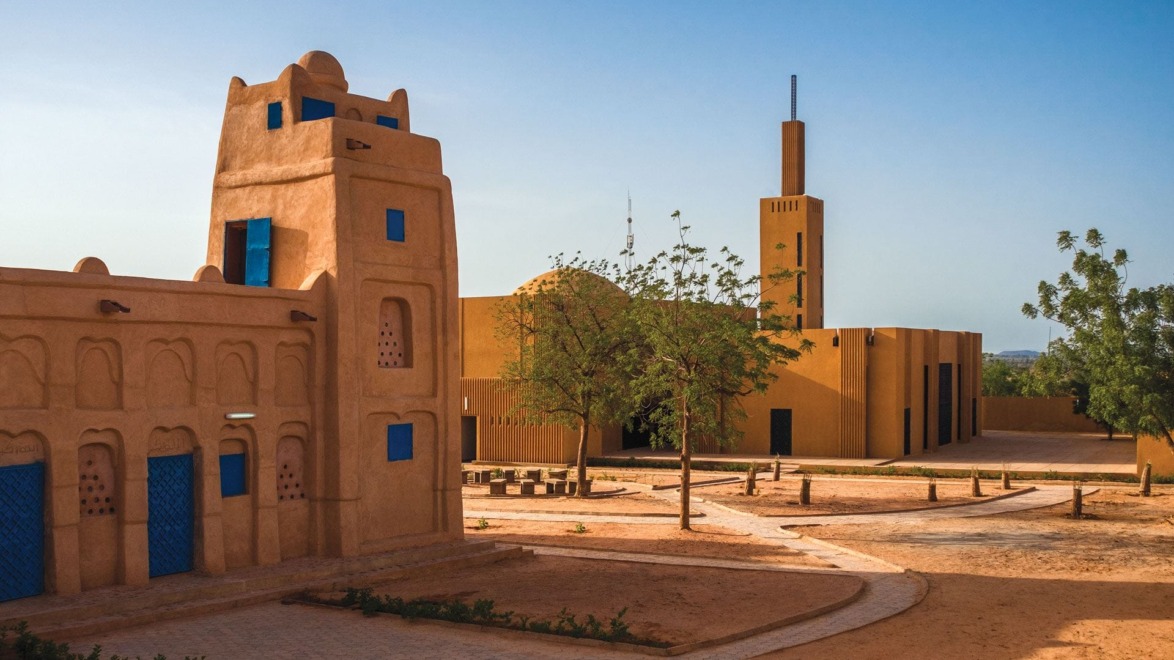 Mud-brick buildings with blue doors under a clear blue sky, surrounded by a sandy landscape and a few trees.