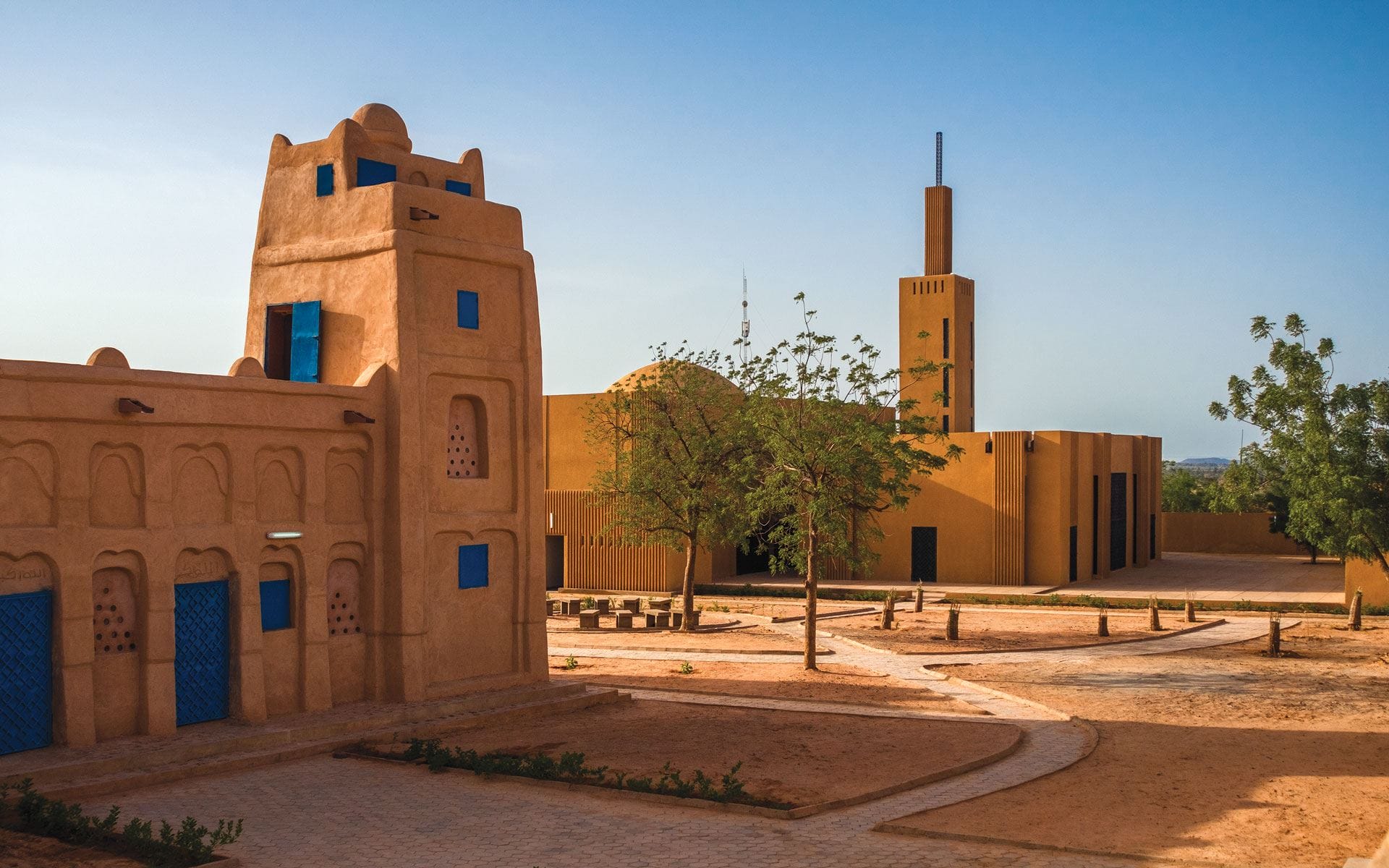 Mud-brick buildings with blue doors under a clear blue sky, surrounded by a sandy landscape and a few trees.