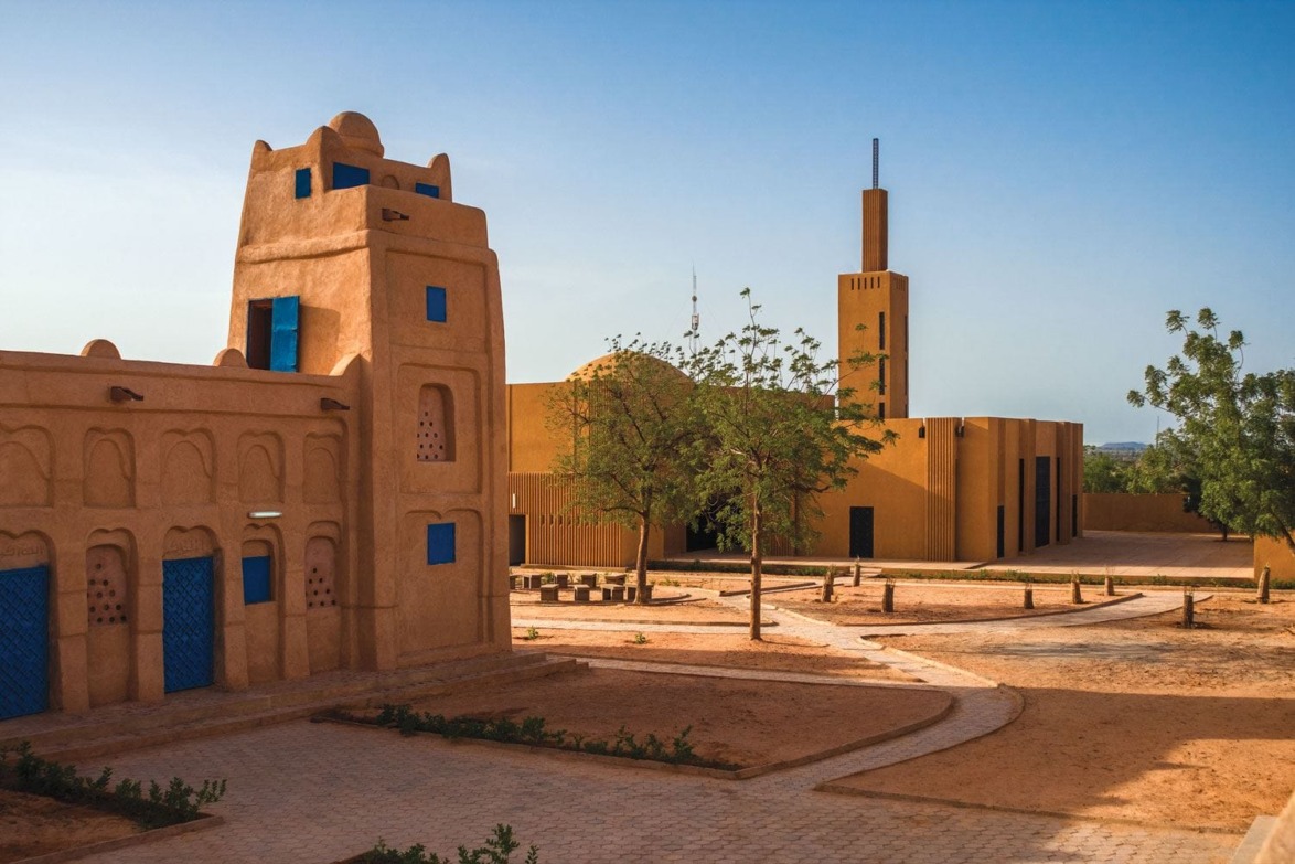 Traditional mud-brick buildings with blue accents in a sunny desert setting featuring trees and a distant tower.