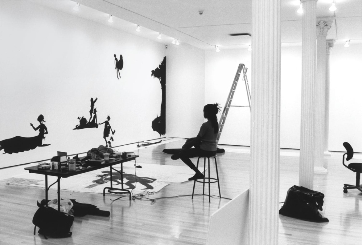 Artist seated in gallery creating silhouette wall art with tools and ladder, surrounded by art supplies and sketches.