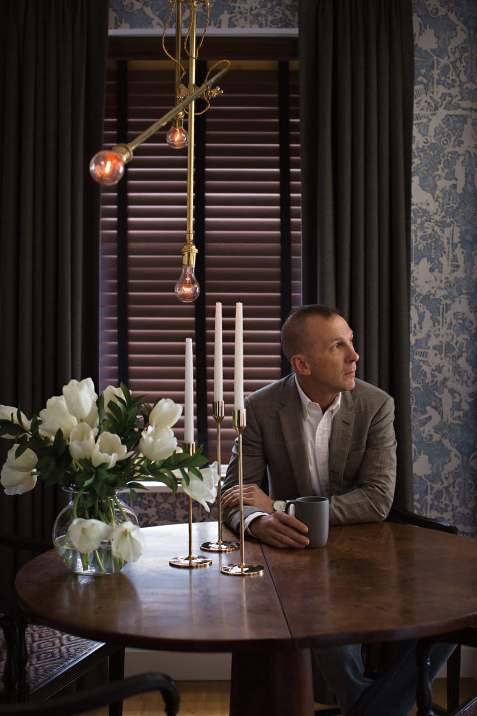 Man sitting at a round wooden table with tulips and candles, holding a mug, looking out a window under hanging bulbs.