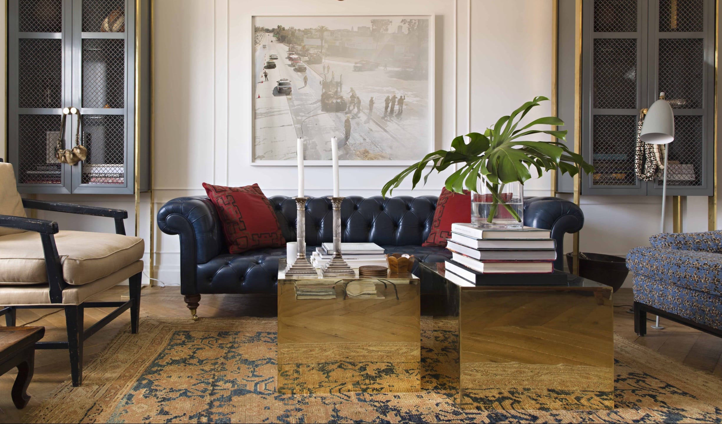 Modern living room with navy sofa, cream armchair, gold coffee tables, and tropical plant. Large artwork on the wall.