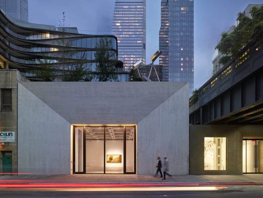 Contemporary art gallery exterior with glass entrance, city buildings in background, and two people walking by at dusk.