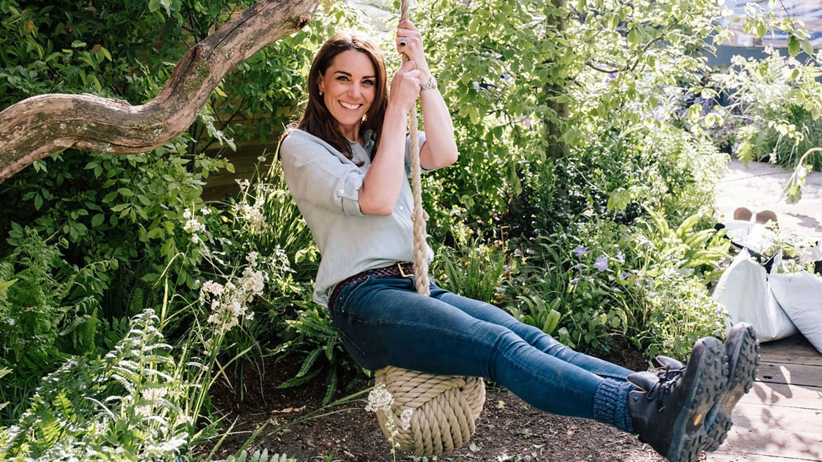 Person sitting on a rope swing in a lush garden, surrounded by green plants and flowering bushes.