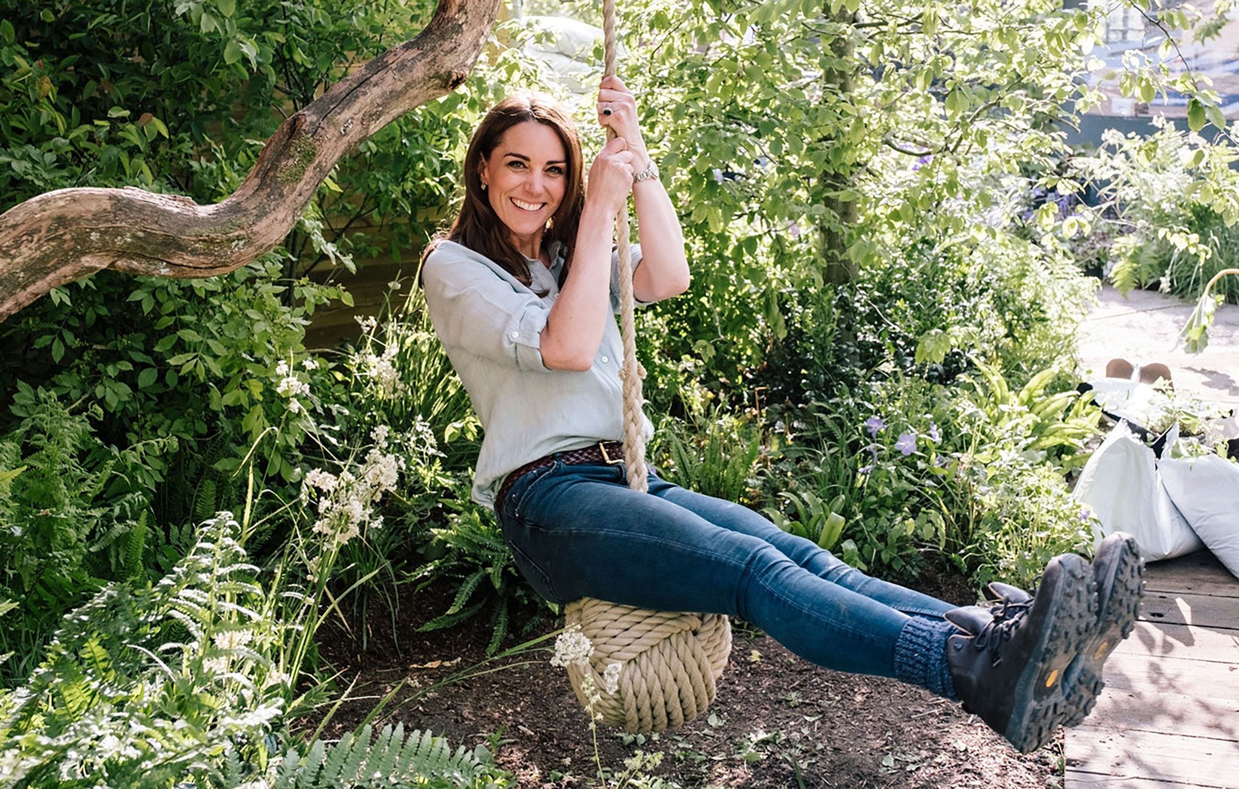 Person sitting on a rope swing in a lush garden, surrounded by green plants and flowering bushes.