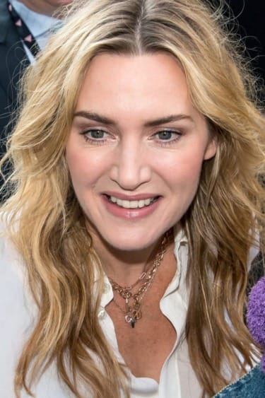 Person with long blonde hair smiles, wearing a white top and necklaces, amid a crowd.