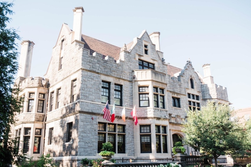 Historic stone mansion with flags and large windows on a sunny day, surrounded by trees and greenery.