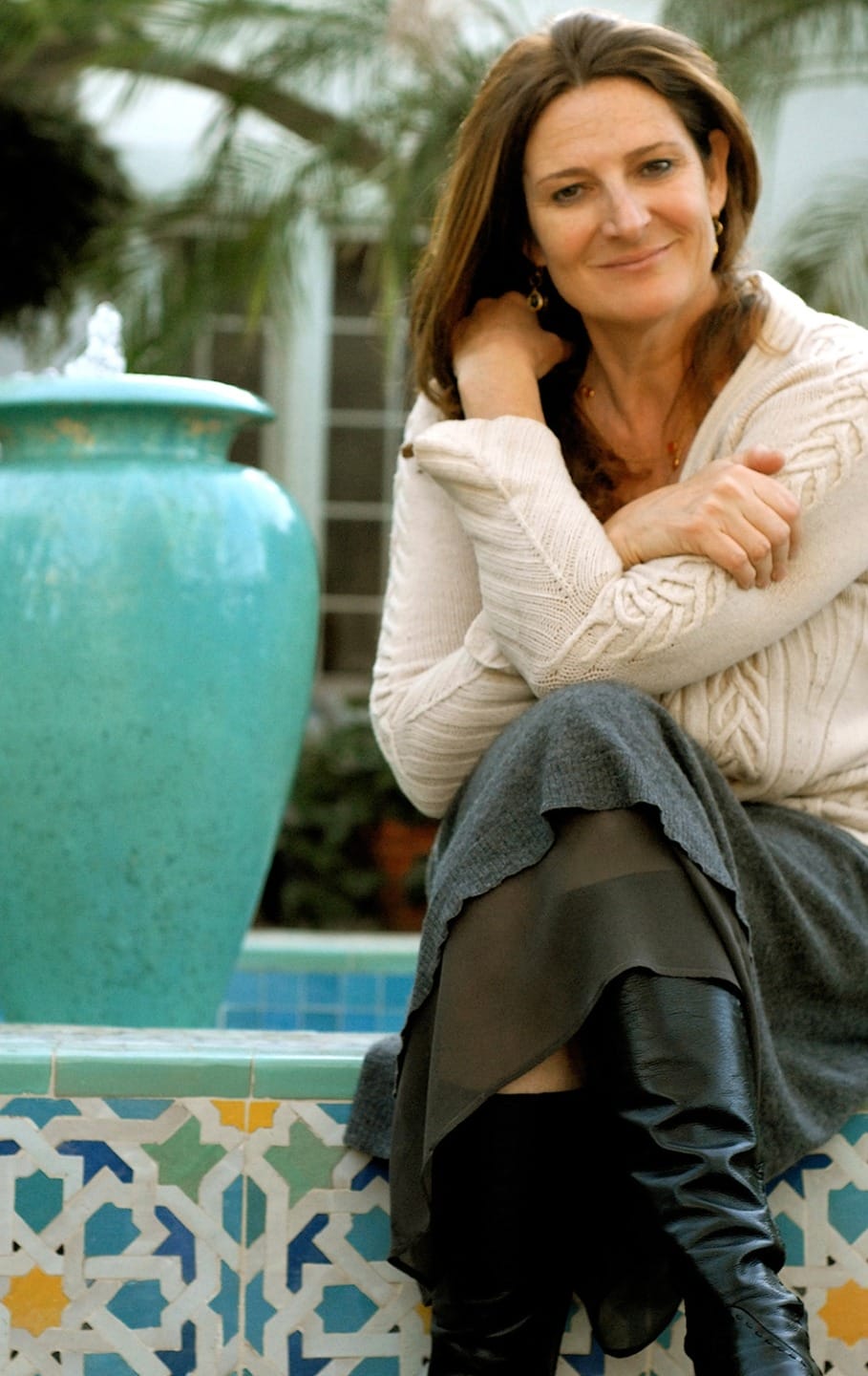A person sitting on a tiled bench near a turquoise vase in a garden setting, wearing a sweater and boots.