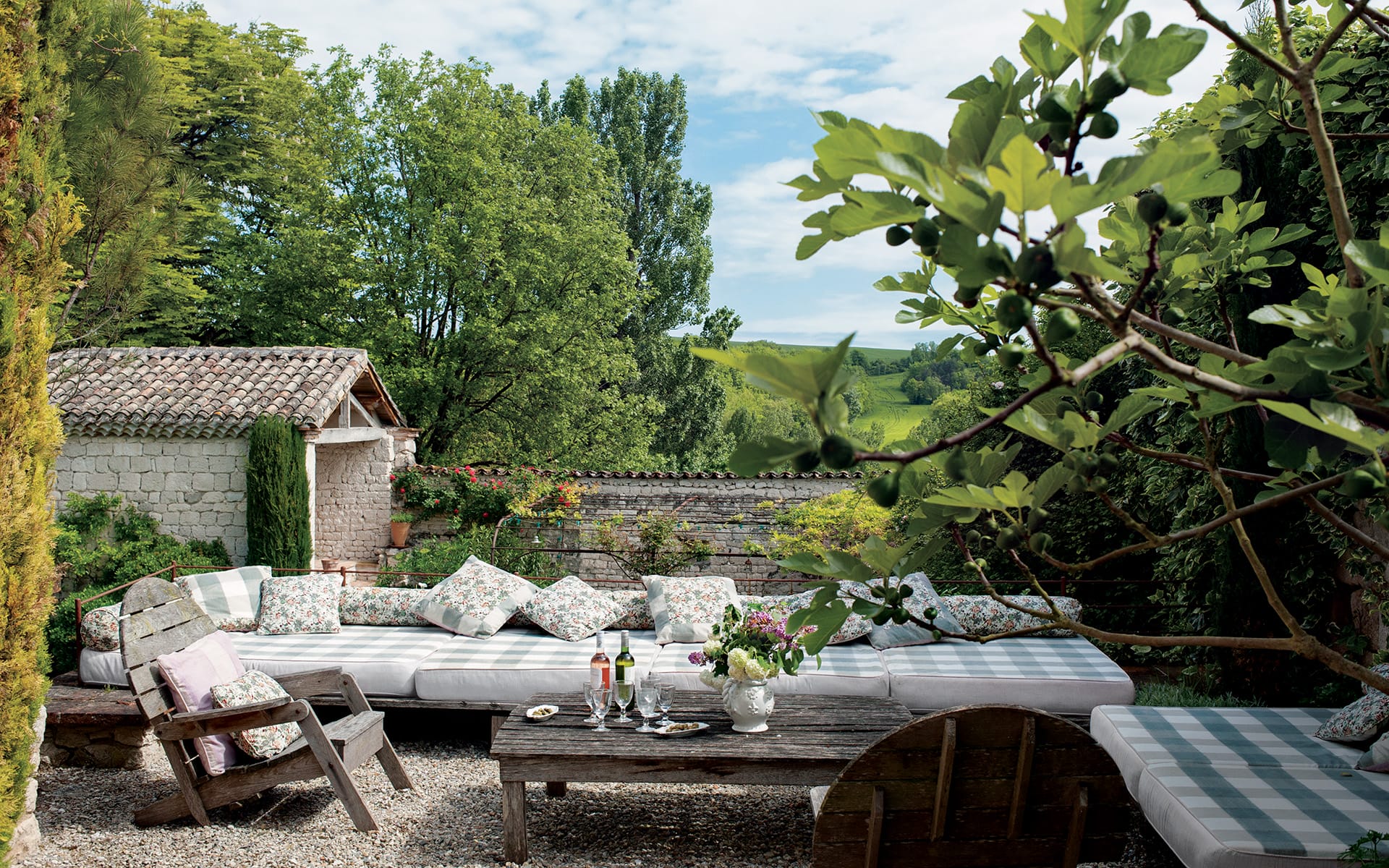 Outdoor patio with cushioned seating, rustic table, wine glasses, and lush greenery overlooking a serene countryside view.