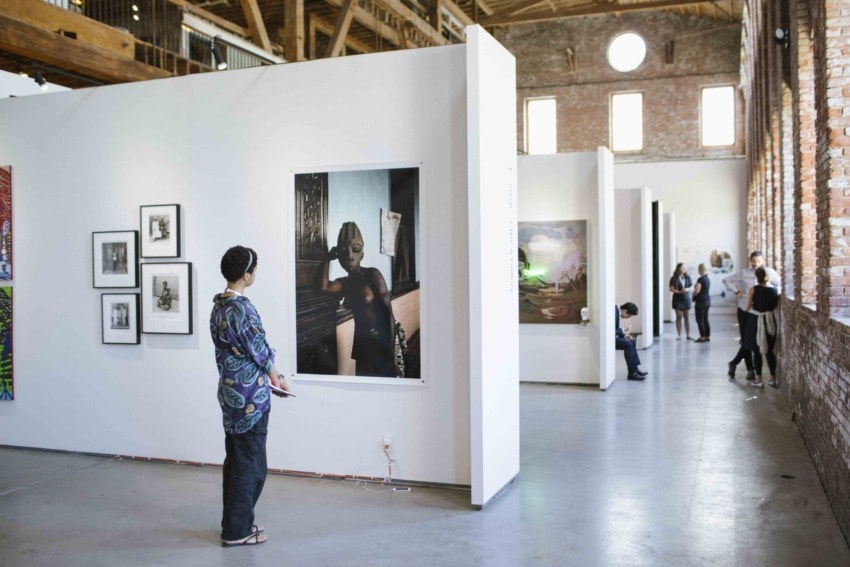 Visitor observing artwork in modern art gallery with brick walls and wooden ceiling beams, other people viewing art in background.
