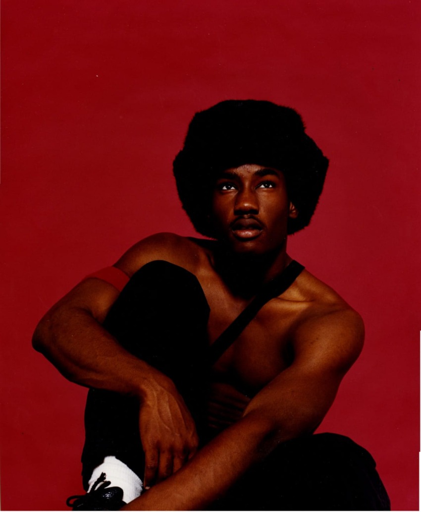 Man sitting shirtless against red background, wearing a large black hat, looking thoughtfully upwards.