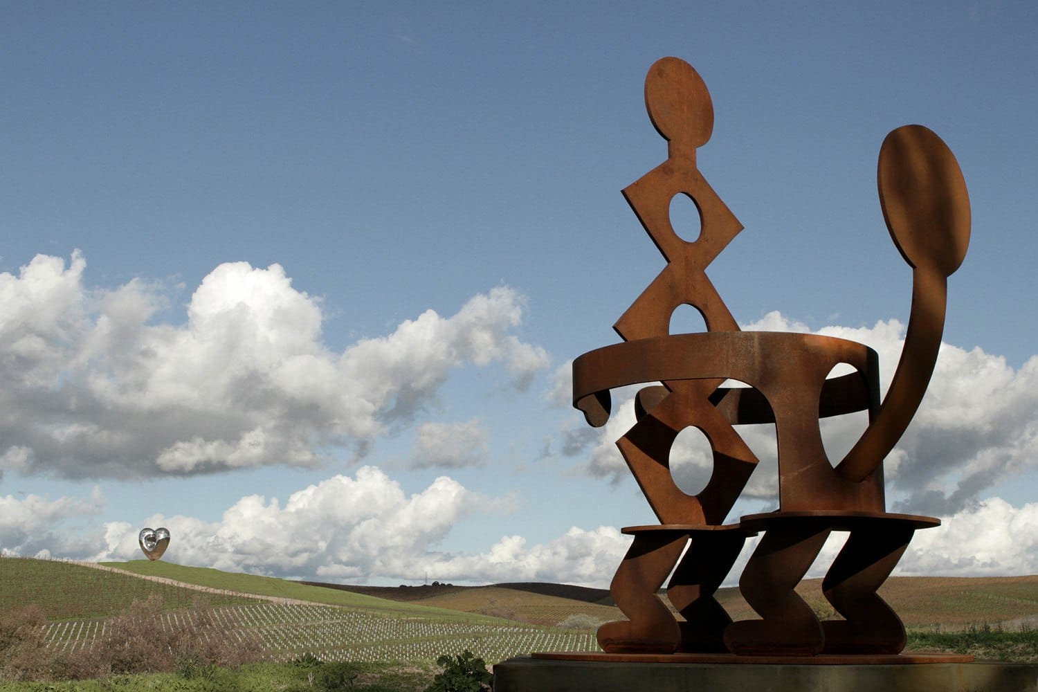 Abstract metal sculptures in a vineyard landscape under a partly cloudy sky.