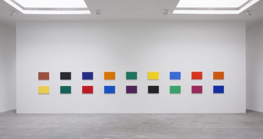 Exhibition room with white walls displaying an array of colorful square paintings in a grid, under large skylights.