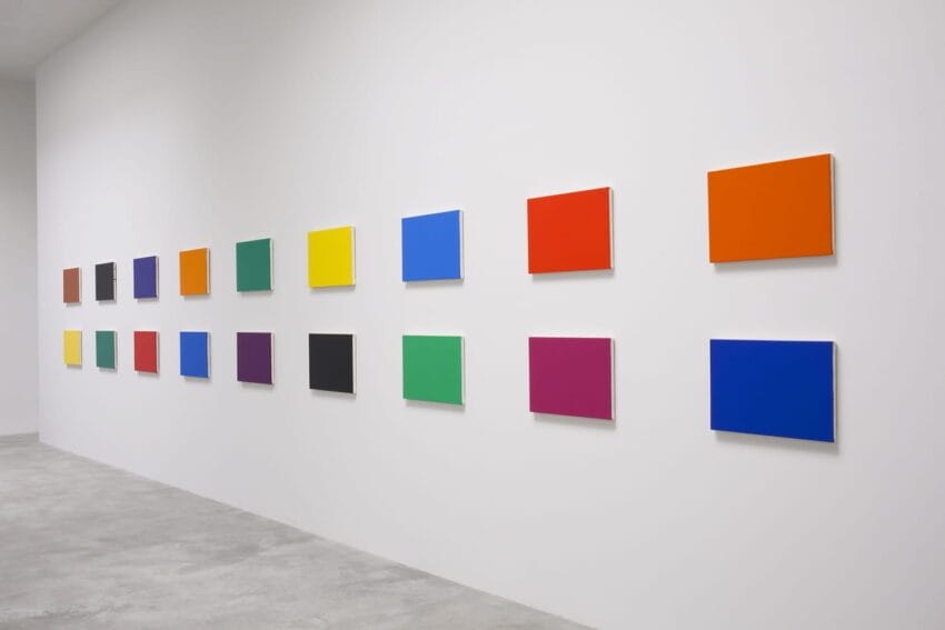 Colorful abstract paintings arranged in a grid on a white gallery wall.