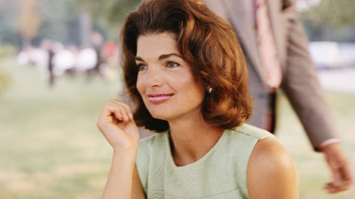 Woman in a green dress sitting outdoors, smiling with a blurred person in the background.